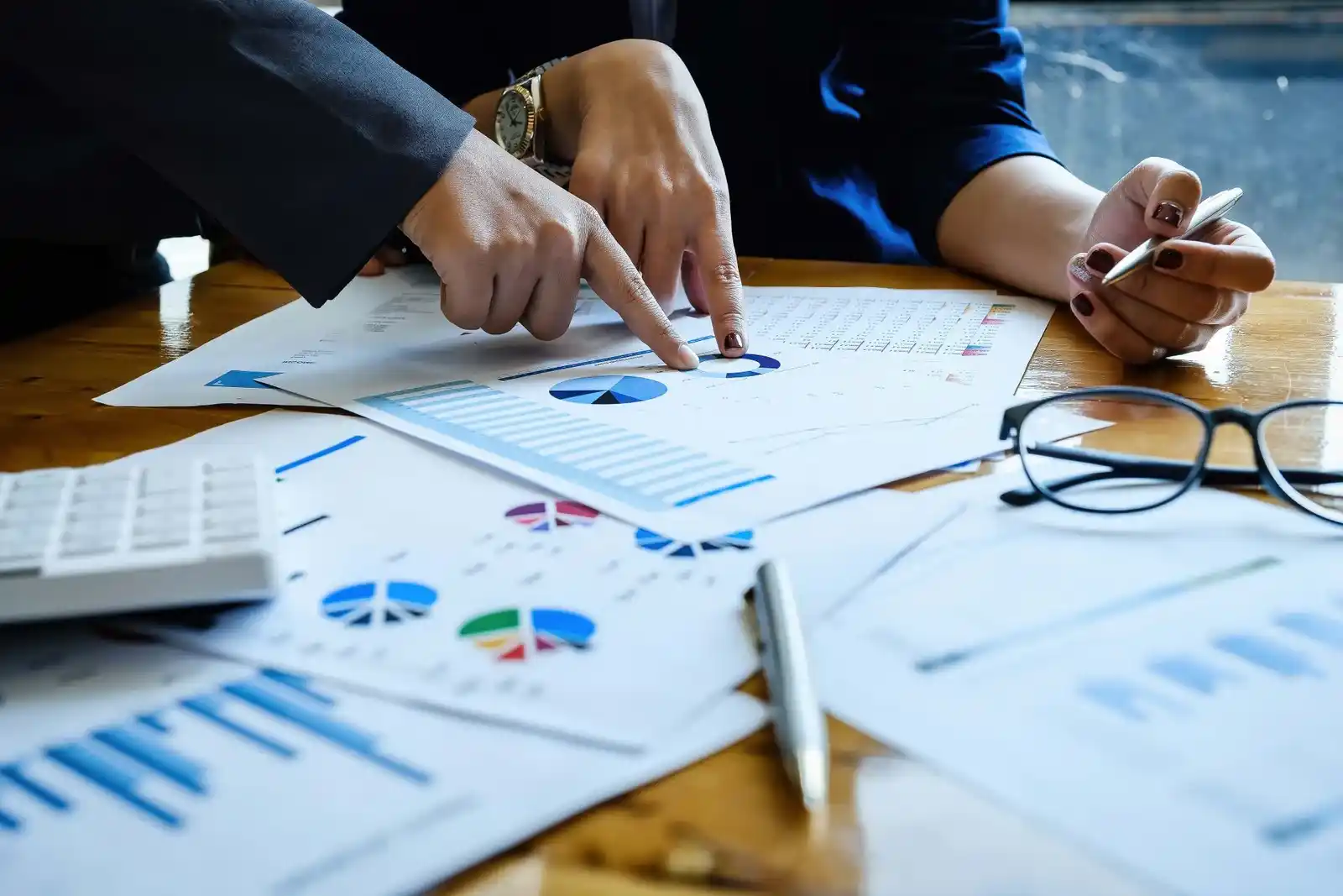 Two people analyze financial charts and data on a wooden table, using a calculator and tax materials related to tax accounting services.